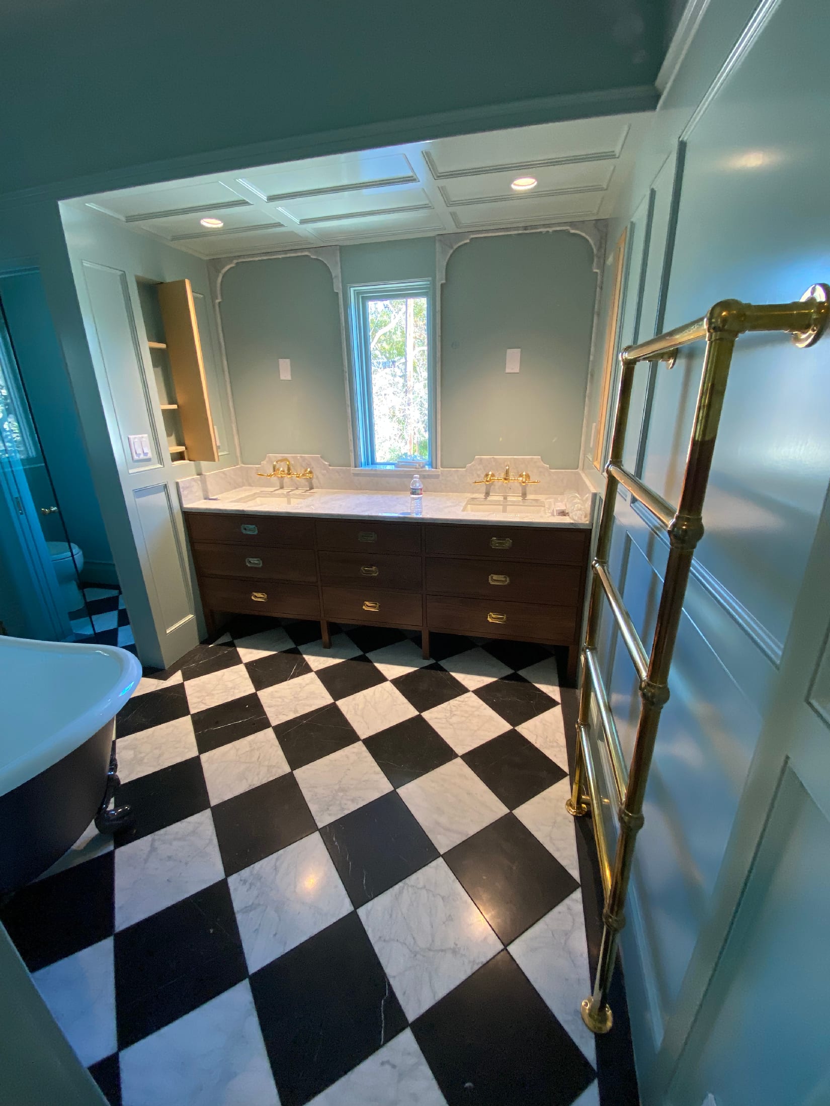 View through a master bath doorway: walnut vanity, marble counters, and a checkered marble floor.