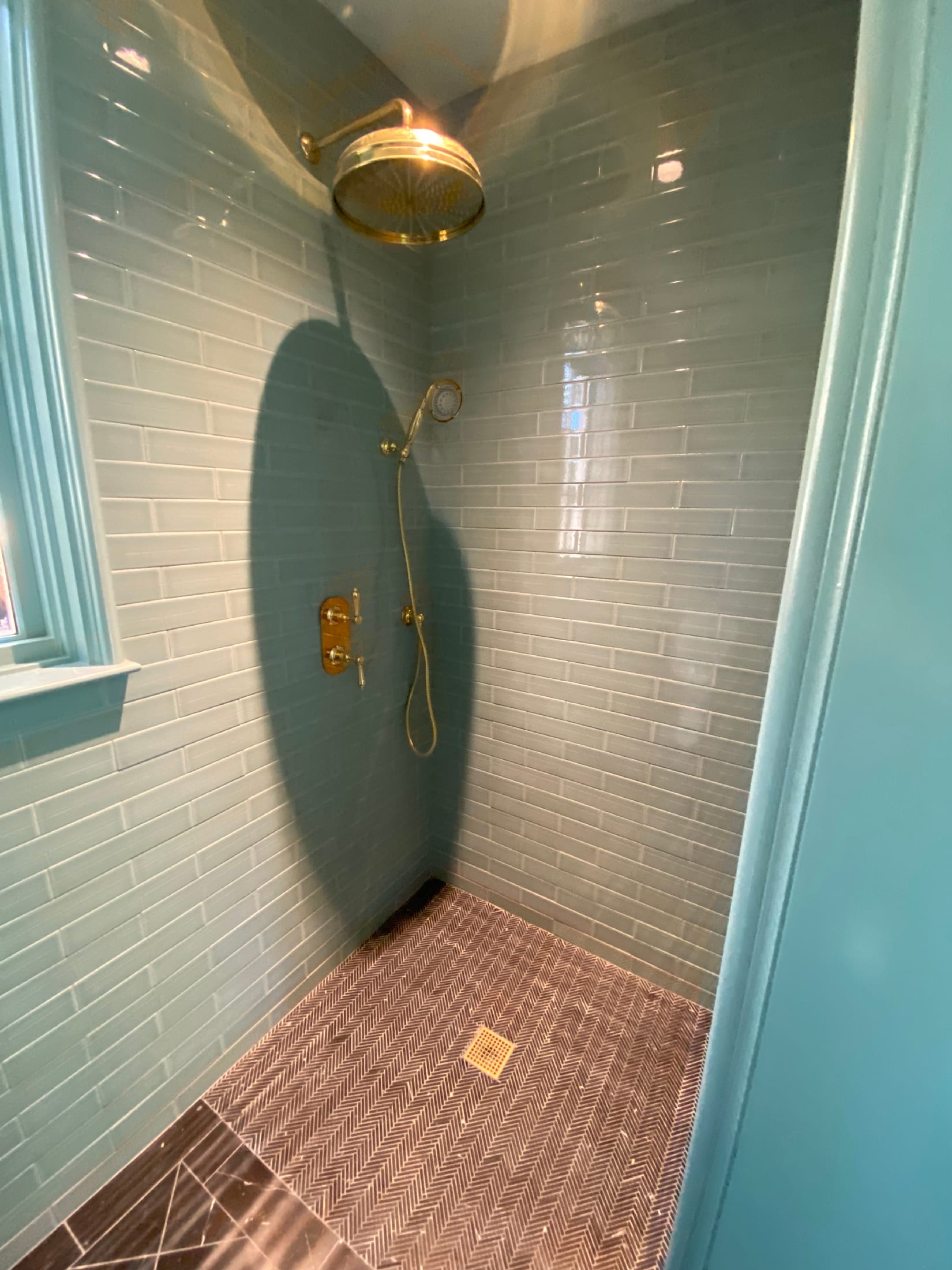 Brass rain shower head and trim against a soft blue glass subway tile shower with a herringbone marble pan.