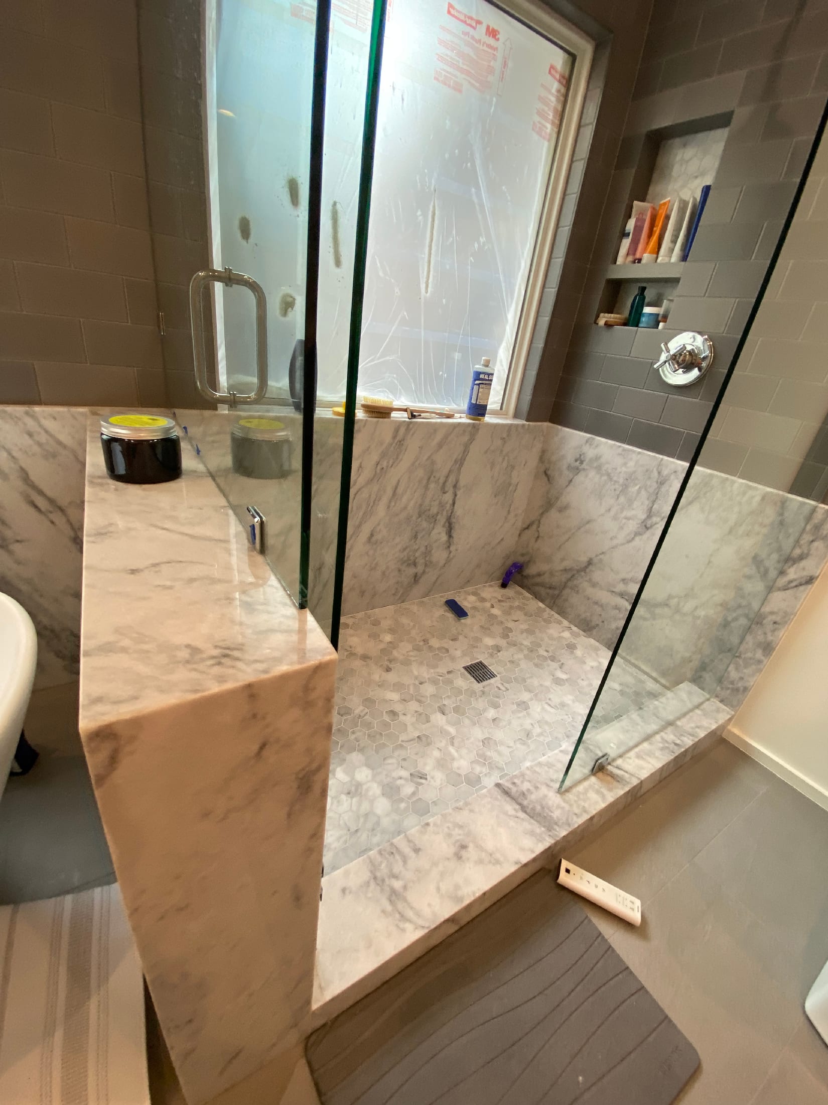 Custom Carrara marble shower bench framed in glass, with gray subway tile walls and a hex marble floor.